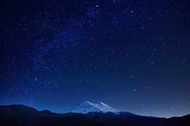 Karla kaplı bir zirve ile nefes kesici bir dağ canlı yıldızlarla dolu berrak bir gece gökyüzüne karşı kurulmuş ve derin mavi tonlardan oluşan zengin bir zemin, sakin bir atmosfer yaratıyor..
