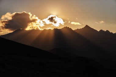 Güneş ışığı bulutların arasından doğar, gün batımında engebeli dağların tepelerine ışınlar saçar, akşam gökyüzünde nefes kesici doğa manzarası yaratır..
