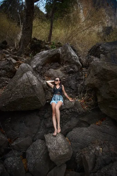 Kadın, suyun kenarındaki büyük kayalara uzanıp güneşin sıcaklığının tadını çıkarıyor. Gösterişli bir kıyafet ve güneş gözlüğü takıyor. Etrafı tropikal bir çevre ve yemyeşil..