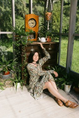 Genç bir kadın, çeşitli bitkilerle çevrili bir serada huzur dolu bir anın tadını çıkarıyor. Güneş ışığı, klasik ahşap bir saatin yanında durduğu sırada huzur dolu bir ortam yayıyor..