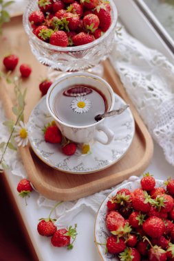 Ahşap bir tepsi iki kase kırmızı çilekle süslenir ve üzerine bir papatya konmuş bir fincan çay ilave edilir. Bu ortam sıcak ve davetkar bir öğleden sonra toplanmasını çağrıştırıyor..