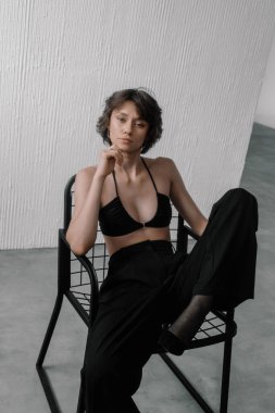 Model, parlak, minimalist stüdyo alanı içinde parlak bir sandalyede oturan siyah bikinili ve bol pantolonlu çarpıcı bir poz sergiliyor. Doğal ışık bölgeyi aydınlatıyor..
