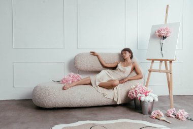 Kadın zarif bir şekilde pembe çiçeklerle çevrili eşsiz bir kanepeye uzanır ve parlak ve şık bir iç mekânda çiçek sanatını sergileyen bir sehpa sergiler..