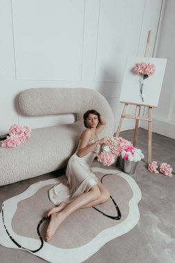 Model, parlak bir stüdyoda yumuşak bir halının üzerinde, pastel çiçekler ve sehpalarla çevrili, yaratıcı bir fotoğraf çekimi sırasında huzurlu bir atmosfer sergiliyor..