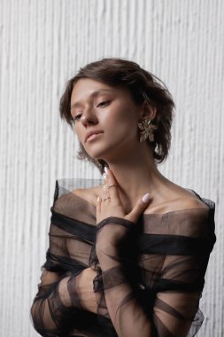 Genç bir kadın eli boynuna yakın poz veriyor, katıksız siyah bir bluz giyiyor. Yumuşak ışıklandırma beyaz bir duvara karşı onun özelliklerini vurgular.