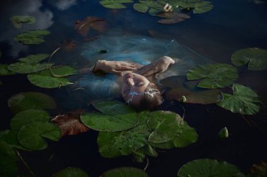 Kadın, etrafını saran nilüferlerle dolu durgun bir göletin üzerinde güzelce dinleniyor. Sakin su mavi gökyüzünü yansıtır, huzurlu bir atmosfer yaratır..
