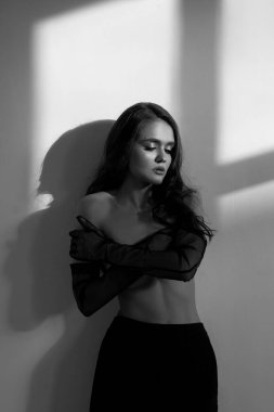 Bir kadın siyah ve beyaz bir ortamda güven içinde poz verir, kolları çapraz ve gölge oynaması ifadesini güçlendirir. Gün ortasında parlak bir ışık pencereden geçer..