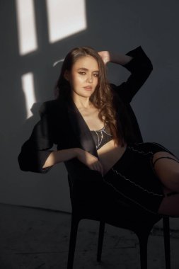 Bir model zarif bir şekilde sandalyede otururken şık bir kıyafet sergiliyor, yumuşak güneş ışığı minimalist stüdyo ortamında ilgi çekici gölgeler bırakıyor..