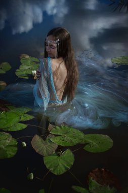 Uzun saçlı mistik karakter zarif bir şekilde sakin sularda oturur, narin kumaşlarla süslenir ve parçalı bulutlu bir gökyüzünün altında yemyeşil ile çevrilidir..