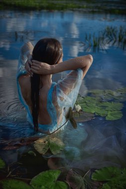 Yarı şeffaf mavi bir elbise giyen kadın saçlarını zarifçe düzeltir. Gecenin erken saatlerindeki yumuşak ışığı yansıtan nilüferlerle dolu sakin suyun içinde dururken..