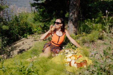 Şık giyinmiş genç bir kadın, sakin bir gölün kenarındaki meyve ve çiçeklerle süslenmiş bir battaniyede dinlenirken içkisini yudumluyor ve ılık güneş ışığında güneşleniyor..