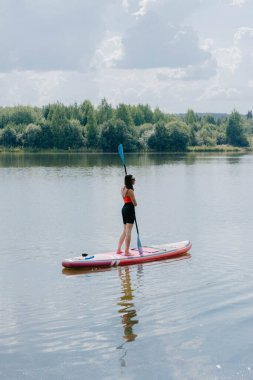Huzurlu bir nehrin ortasında bir kadın kürek tahtasının üzerinde duruyor. Güneş yemyeşil ağaçların üzerinde ışıl ışıl parlıyor suları çevreleyen, sakin bir atmosfer yaratıyor..