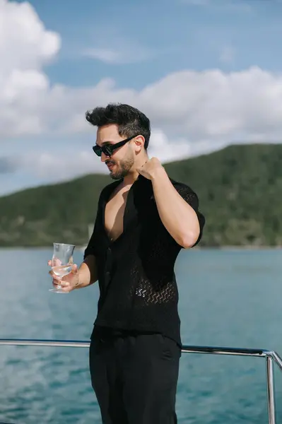 Siyah elbiseli genç bir adam bir tekne partisinde bir bardak şampanyayla eğleniyor.