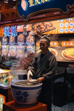 Şangay, Çin; 14 Ekim 2024: Çinli bir adam Nanjing yolunda popüler gıda pazarında erişte pişiriyor; müşterileri dükkana çekmek için bir mikrofon kullanıyor.