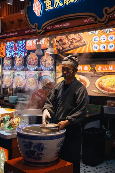 Şangay, Çin; 14 Ekim 2024: Çinli bir adam Nanjing yolunda popüler gıda pazarında erişte pişiriyor; müşterileri dükkana çekmek için bir mikrofon kullanıyor.