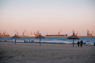 Qinhuangdao, China; October 19 2024: Tourist beach with people walking and petrochemical factories with oil and gas transportation boats on the shore clipart