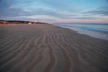 Şafak vakti dalgalı kumların üzerinde dalgalı dalgalar, uzak binalar ve pastel gökyüzü; Rota, Cadiz, İspanya