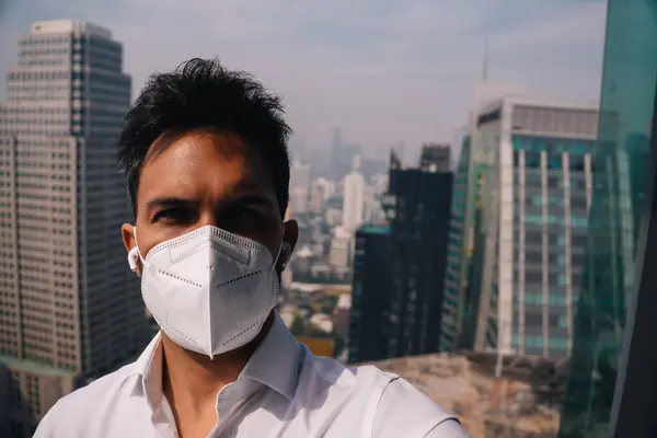 N95 yüz maskesi ve kablosuz kulaklık takan bir iş adamı arka planda şehir manzarası olan bir çatıda selfie çekiyor.