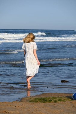 Hafif beyaz elbiseli romantik sarışın bayan sıcak ve güneşli bir günde sahil yürüyüşünün tadını çıkarıyor. İnanılmaz Kuzey Denizi 