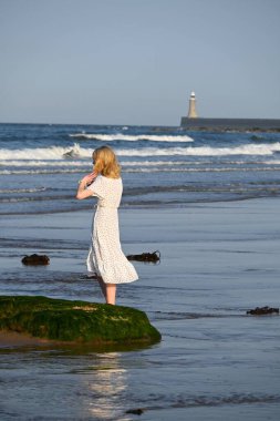 Young romantic blonde lady  with a light weighted white dress on enjoys her beechwalk on a hot sunny day. Incredible North Sea  clipart