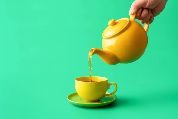 Woman\'s hand pour tea from yellow jug in a cup, minimalist on a green table. Hot healthy drink, mint tea in a yellow-colored cup.