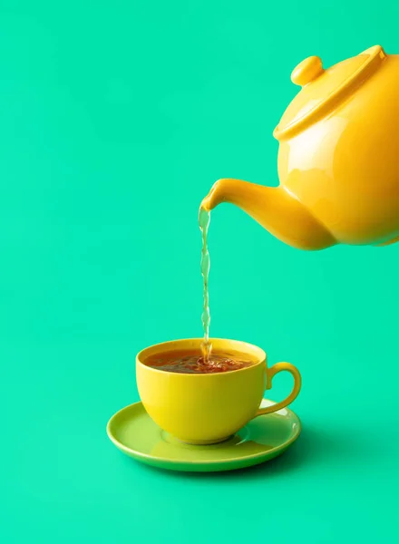 Pouring tea from a ceramic teapot in a cup, minimalist on a green table. Hot mint tea in a yellow colored cup isolated on a green background