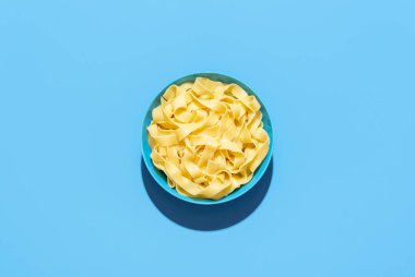 Üstünde pappardelle makarnası olan bir kâse, mavi bir masada minimalist. Canlı renkli arka planda parlak ışıkta sade erişte.