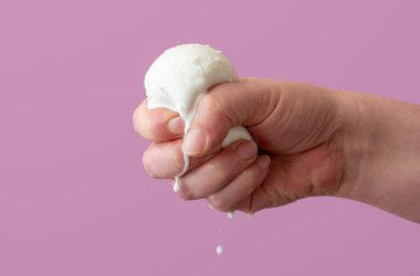 A woman's hand is squeezing a ball of mozzarella cheese, minimalist on a purple background. Buffalo milk mozzarella in a woman's hand.