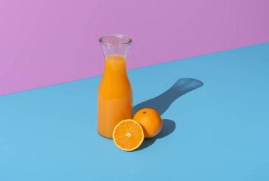Freshly squeezed orange juice in a glass carafe minimalist on a blue table. Oranges and orange juice in bright light on a vibrant colored background.