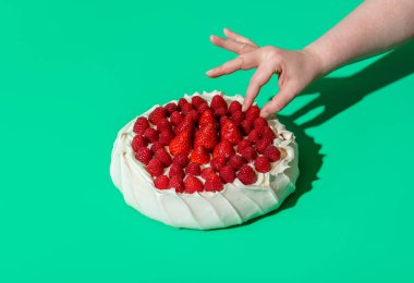 Woman's hand grabbing a raspberry from a pavlova cake, minimalist on a green background. Homemade pavlova cake with whipped cream filling, strawberries, and raspberries on top of it.