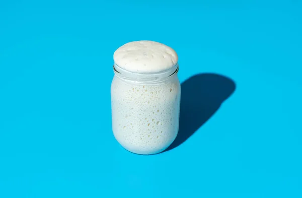 Jar with an active sourdough starter in bright light minimalist on a blue table. Healthy home baking with sourdough starter.
