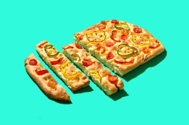 Dilimlenmiş Focaccia parlak ışıkta yeşil bir masada. Çeri domatesli ve kırmızı biberli ev yapımı vegan Focaccia.