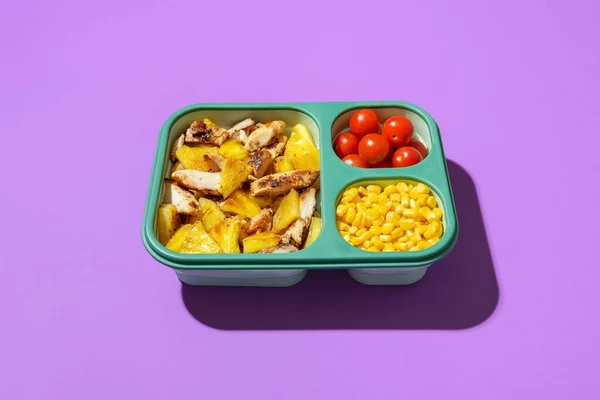Chicken and pineapple salad in a silicone lunch box, minimalist on a purple table. Sweet and sour dish, salad with roasted pineapple, chicken, corn and cherry tomatoes.