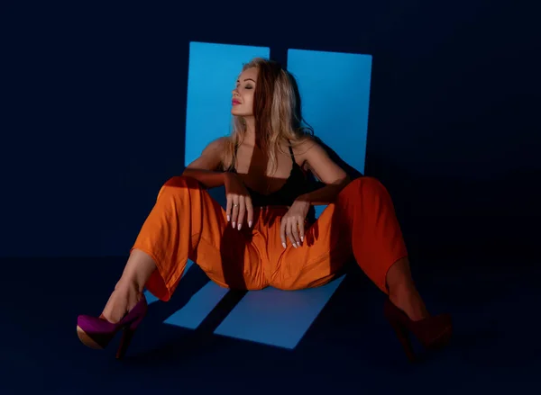 stock image Blonde woman in window light sitting and leaning on a blue wall. Mature woman in orange pants relaxing in a bright window light