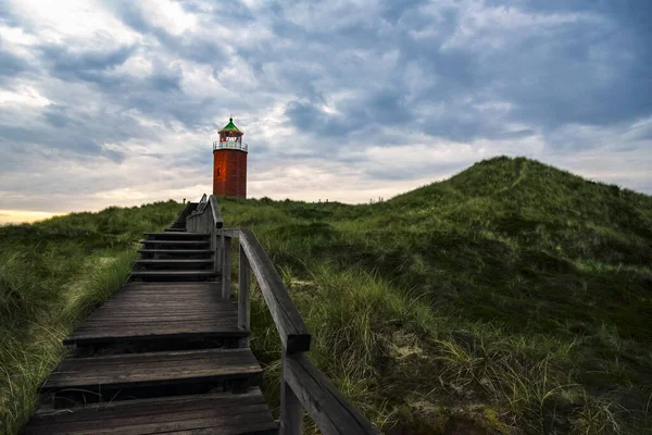 stock image Evening landscape with wooden stairs over marram grass hill, toward red lighthouse, on Sylt island, Germany