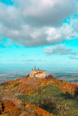 Güneşli bir günde tepedeki Hohenzollern kalesi, sonbahar ormanıyla çevrili, mavi bir gökyüzünün altında.