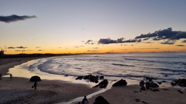 Okyanus kıyı şeridini turlarken ufukta gün batımları beliriyor. Sörfçülerin siluetleri Tge Sahili 'ni terk ederken görülebilir. Körfezin karşısındaki şehir, güneş ufukta parlak bir portakal gibi parlarken uzaktan görülebilir..
