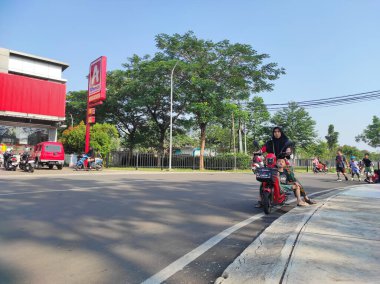 Ciledig, Endonezya - 15 Eylül 2024: annesi ve küçük oğlu kırmızı elektrikli scooter kullanıyor. Bir Alfamidi mağazasının yanındaki yol kenarında park halindeler. Arka planda birkaç motosiklet ve araba var.