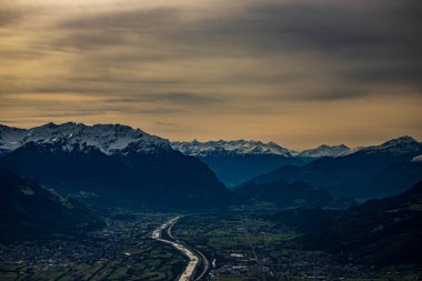 Bulutlu bir gökyüzünün altında yeşilliklerle çevrili güzel bir dağ görüntüsü.