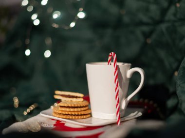 Kırmızı ve beyaz çizgili bir pipetle beyaz bir kupa bir tabakta kurabiyeyle oturuyor. Sahne rahat ve neşeli bir atmosferde geçiyor. Arka planda Noel ışıkları var.