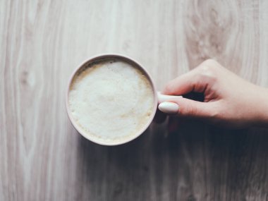 Tahta bir masada bir fincan kahve tutan bir el. Kahve beyaz ve köpüklü, bardak da pembe. Rahatlama ve rahatlama kavramı
