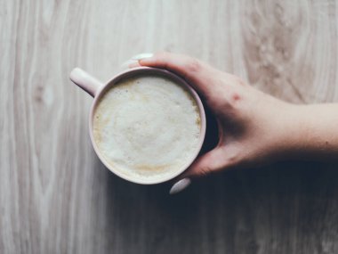 Tahta bir masada bir fincan kahve tutan bir el. Kahve beyaz ve köpüklü, bardak da pembe. Rahatlama ve rahatlama kavramı