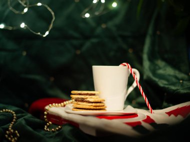 Kırmızı ve beyaz çizgili bir pipetle beyaz bir kupa bir tabakta kurabiyeyle oturuyor. Sahne rahat ve neşeli bir atmosferde geçiyor. Arka planda Noel ışıkları var.