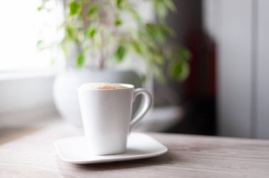Beyaz fincan tabağı olan beyaz bir kahve fincanı ahşap bir masada oturur. Bardak kahve ile dolu ve arka planda bir bitki var.