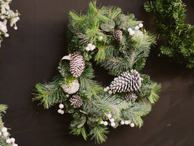 Çam kozalaklarından yapılmış güzel bir çelenk ve duvarda beyaz böğürtlenler asılı. Çam kozalakları çelengin ortasına yerleştirilmiş, bazıları birbiriyle örtüşüyor.