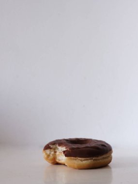 Çikolata kaplı donuttan bir ısırık alınmış. Çörek beyaz bir masada oturuyor.