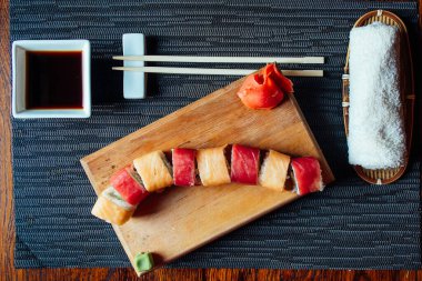 Üzerinde kırmızı ve sarı şerit olan suşi rulosu tahta bir tahtanın üzerinde duruyor. Suşi rulosuna bir kase sos ve yemek çubukları eşlik ediyor. Hoşgörü ve keyif kavramı