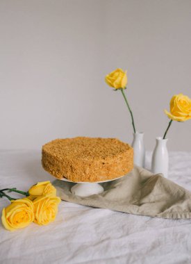Masadaki beyaz bir tabakta duran bir pasta ve arka planda sarı güller var.