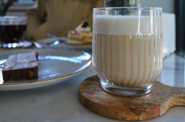 Samimi bir kafede mermer bir masada servis edilen, süt köpüklü, temiz bir bardak latte. Arka planda, bir tatlı barı ve katmanlı pasta kısmen görülebilir, sıcak ve davetkar bir kahvaltı atmosferi yaratıyor.