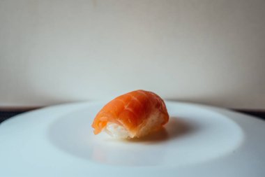 Bir parça suşi beyaz bir tabağın üzerinde duruyor. Suşi ikiye bölündü ve görüntünün ana odağı o. Tabak temiz ve dağınık, bu da suşinin ana denek olarak göze çarpmasına olanak sağlıyor.